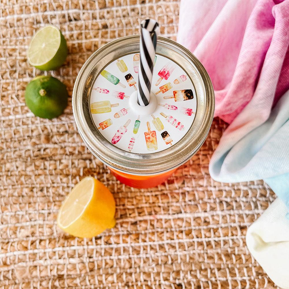 Mason jar straw lid with silver ring and a white sticker that has popsicles in different sizes and colors in print. Photographed as a flat lay in a weave mat with a scarf and some lemon slices.
