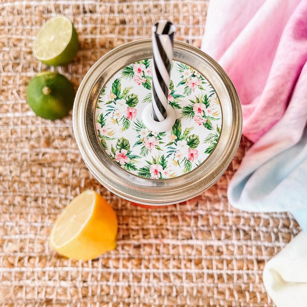 Mason jar straw lid with silver ring and a white sticker that has palm leaves and hibiscus flowers in print. Photographed as a flat lay in a weave mat with a scarf and some lemon slices.
