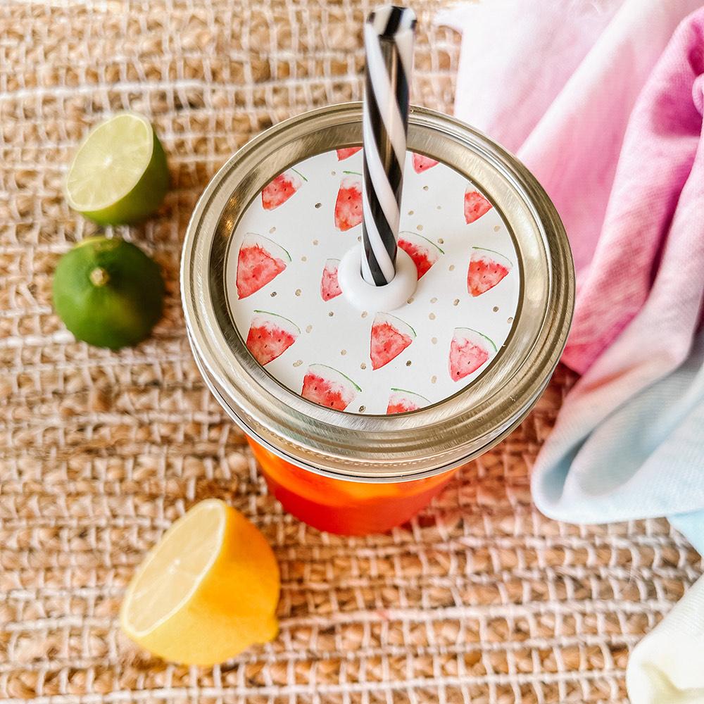 Mason jar straw lid with silver ring and a white sticker that has watermelon slices in print. Photographed as a flat lay in a weave mat with a scarf and some lemon slices.