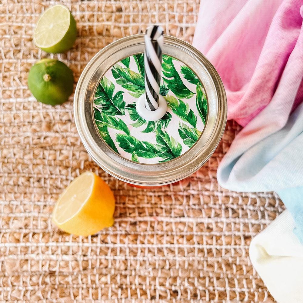Mason jar straw lid with silver ring and a white sticker that has palm leaves in print. Photographed as a flat lay in a weave mat with a scarf and some lemon slices.