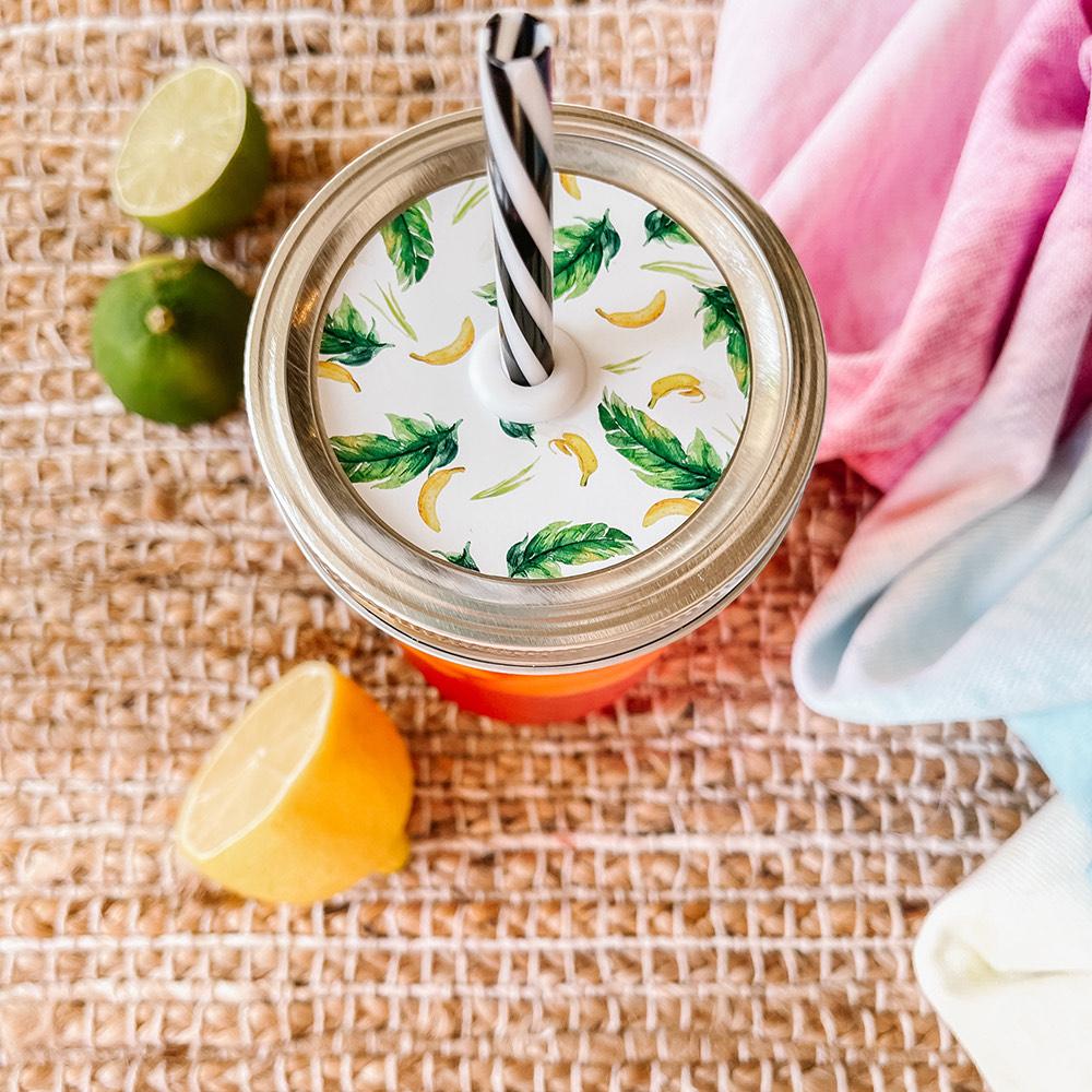 Mason jar straw lid with silver ring and a white sticker with palm leaves and bananas in print. Photographed as a flat lay in a weave mat with a scarf and some lemon slices.