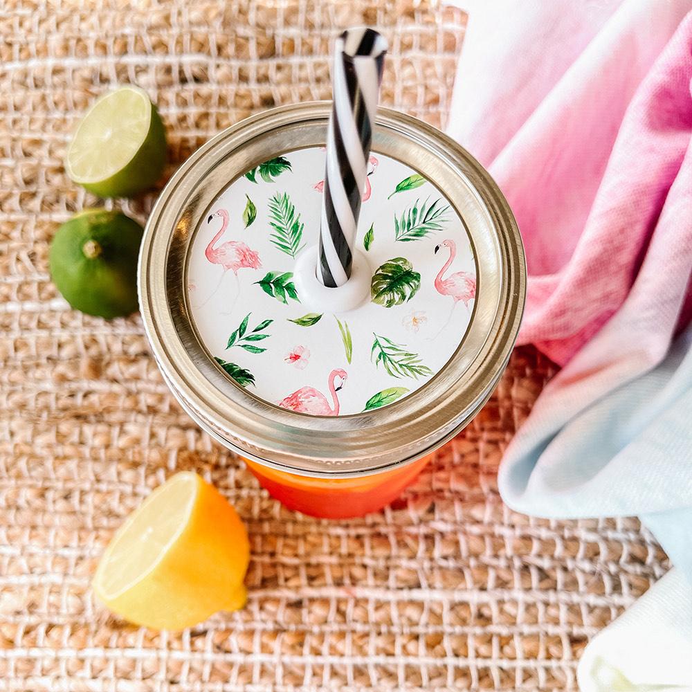 Mason jar straw lid with silver ring and a white sticker that has flamingos, hibiscus flowers, and palm leaves in print. Photographed as a flat lay in a weave mat with a scarf and some lemon slices.