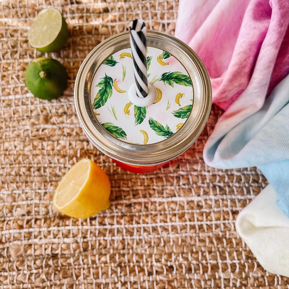 Mason jar straw lid with silver ring and a white sticker that has bananas and palm leaves in print. Photographed as a flat lay in a weave mat with a scarf and some lemon slices.