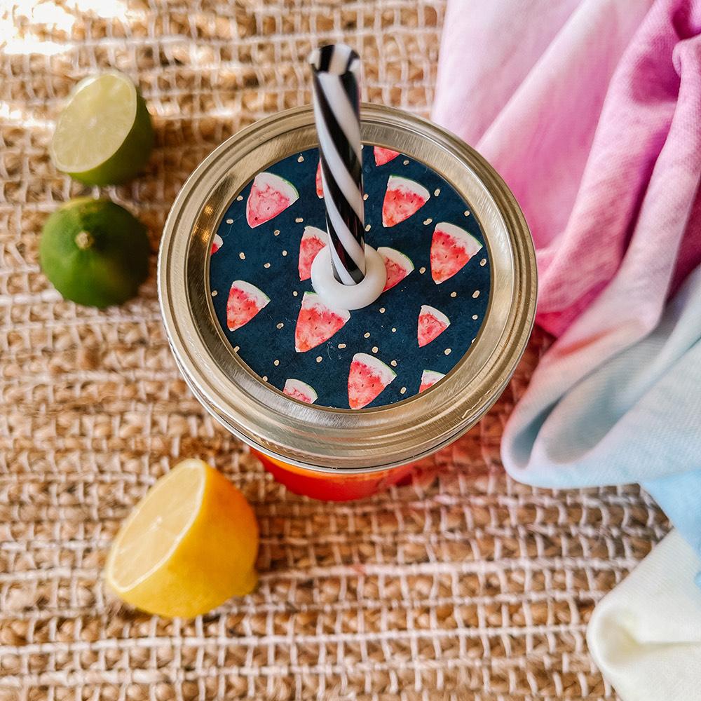 Mason jar straw lid with silver ring and a navy blue sticker that has watermelon slices in print. Photographed as a flat lay in a weave mat with a scarf and some lemon slices.