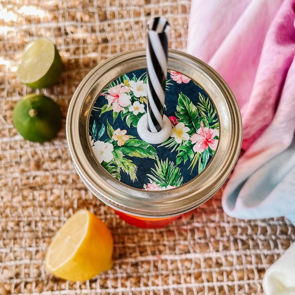 Mason jar straw lid with silver ring and a navy blue sticker that has hibiscus flowers and palm leaves in print. Photographed as a flat lay in a weave mat with a scarf and some lemon slices.