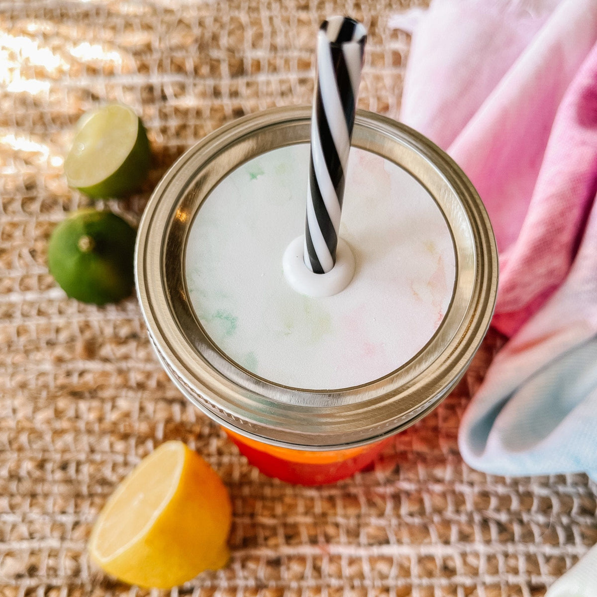 Mason jar straw lid with silver ring and a white sticker with streaks of colors of the beach and sand. Photographed as a flat lay in a weave mat with a scarf and some lemon slices.