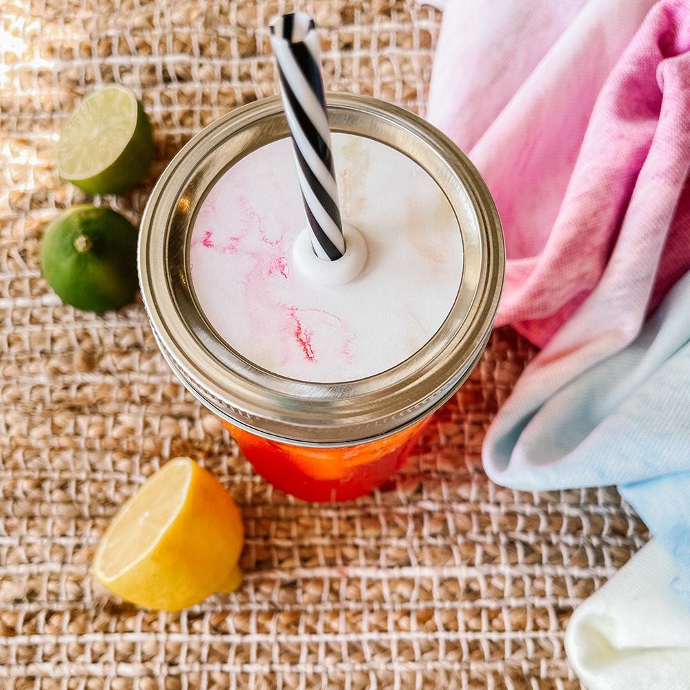 Mason jar straw lid with silver ring and a white sticker with streaks of different shades of pink color similar to a sherbet. Photographed as a flat lay in a weave mat with a scarf and some lemon slices.