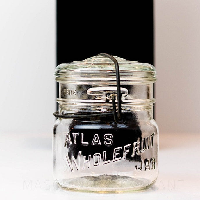 A close up of a Vintage 1940s Atlas Wholefruit wide mouth wire bail mason jar with Atlas whole fruit jar engraved in it against a white background