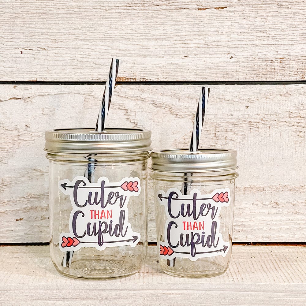 A Regular mouth half-pint (8 oz) and a Wide mouth pint (16 oz) mason jar tumbler with a black and white stripes straw and a text that says &quot;Cuter than Cupid&quot; against a white outline and shown on top of a counter. It has one arrow above the word cute and another below the word cupid. The arrow has three little pink hearts on its tails
