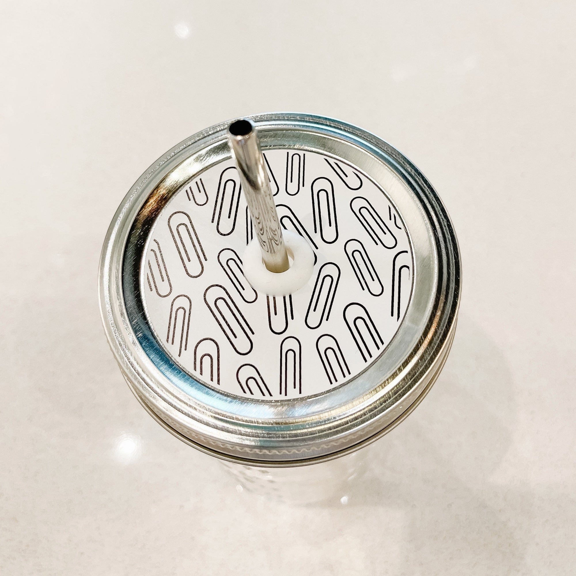 Silver lid with a paper clips on white patterned mason jar straw lid against a white background.