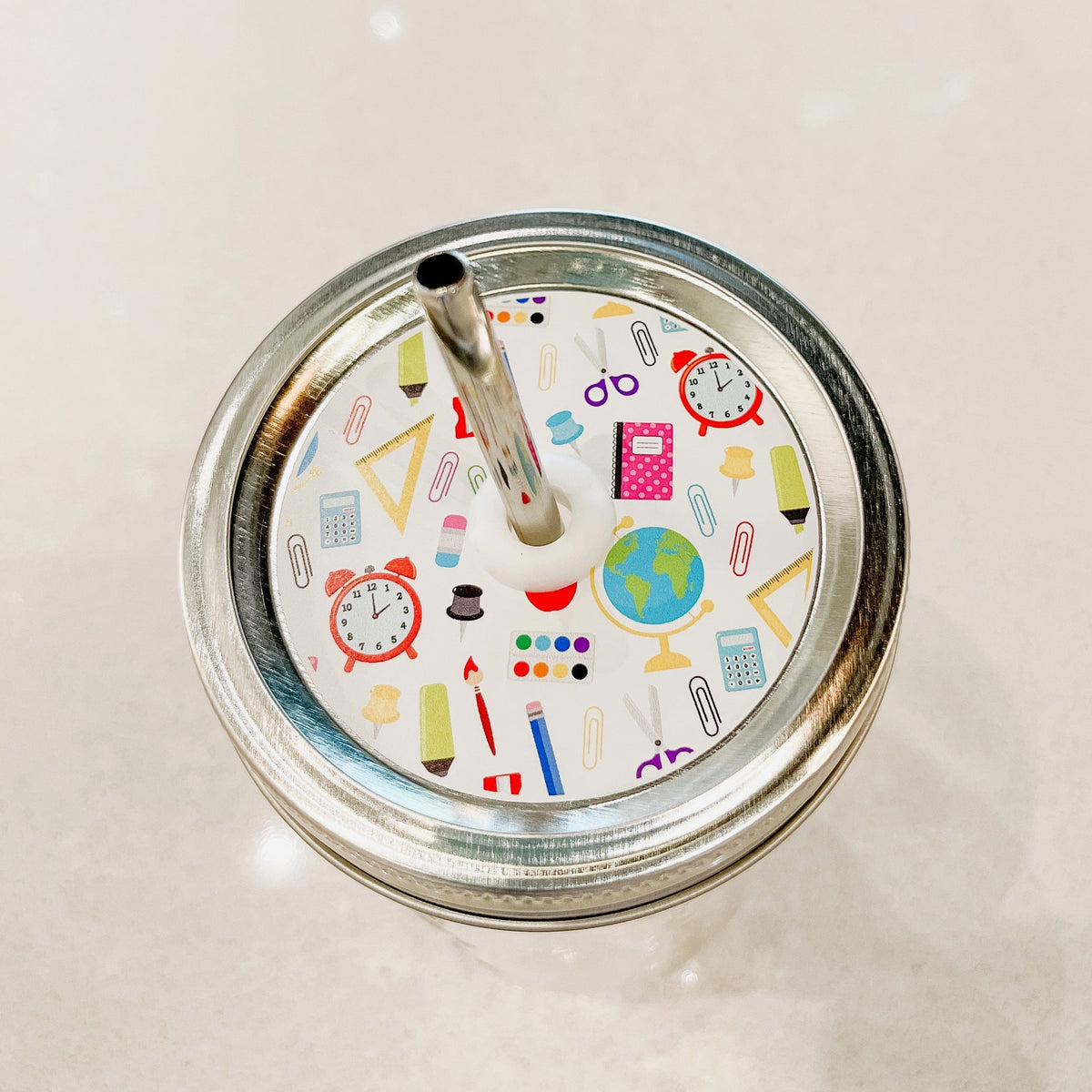 Silver lid with a school supplies patterned mason jar straw lid against a white background.