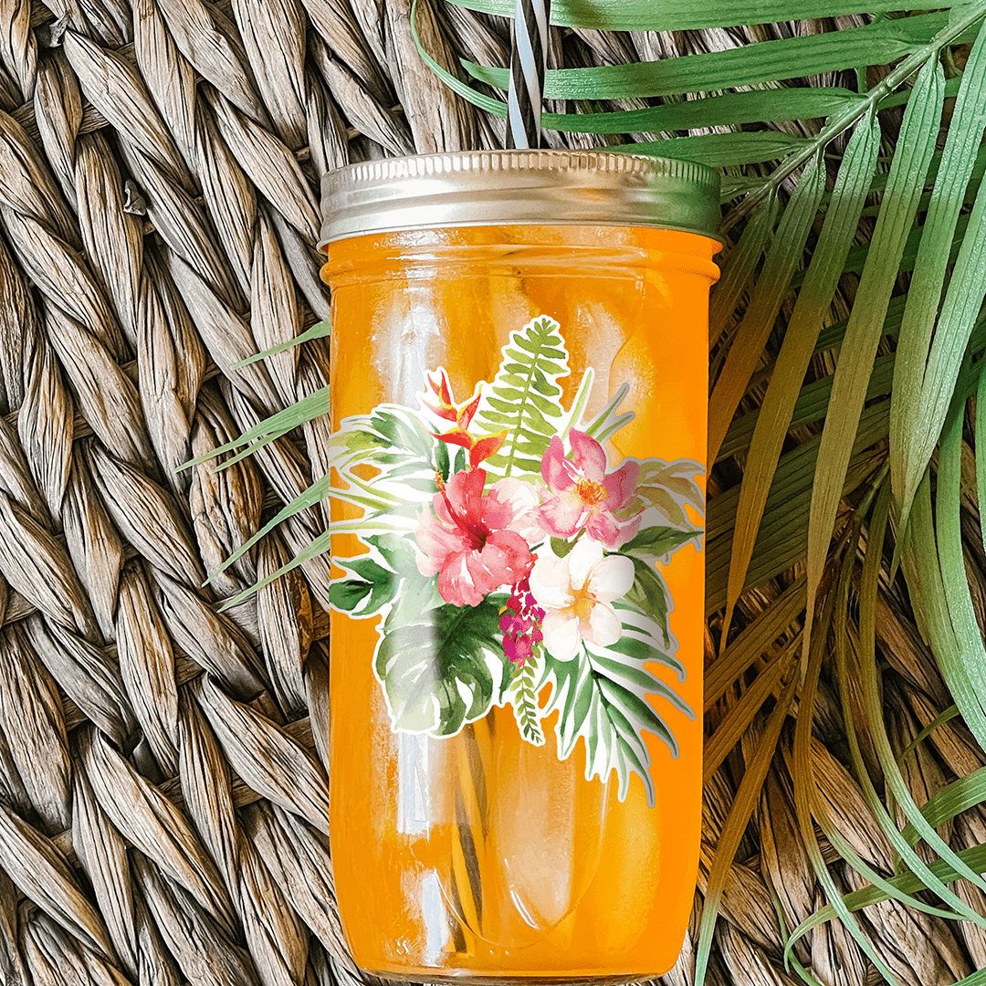 Tumbler with an orange drink and a sticker that has white and pink hibiscus flowers and palm leaves as graphic details. Photographed as a flat lay in a weave mat with palm leaves.