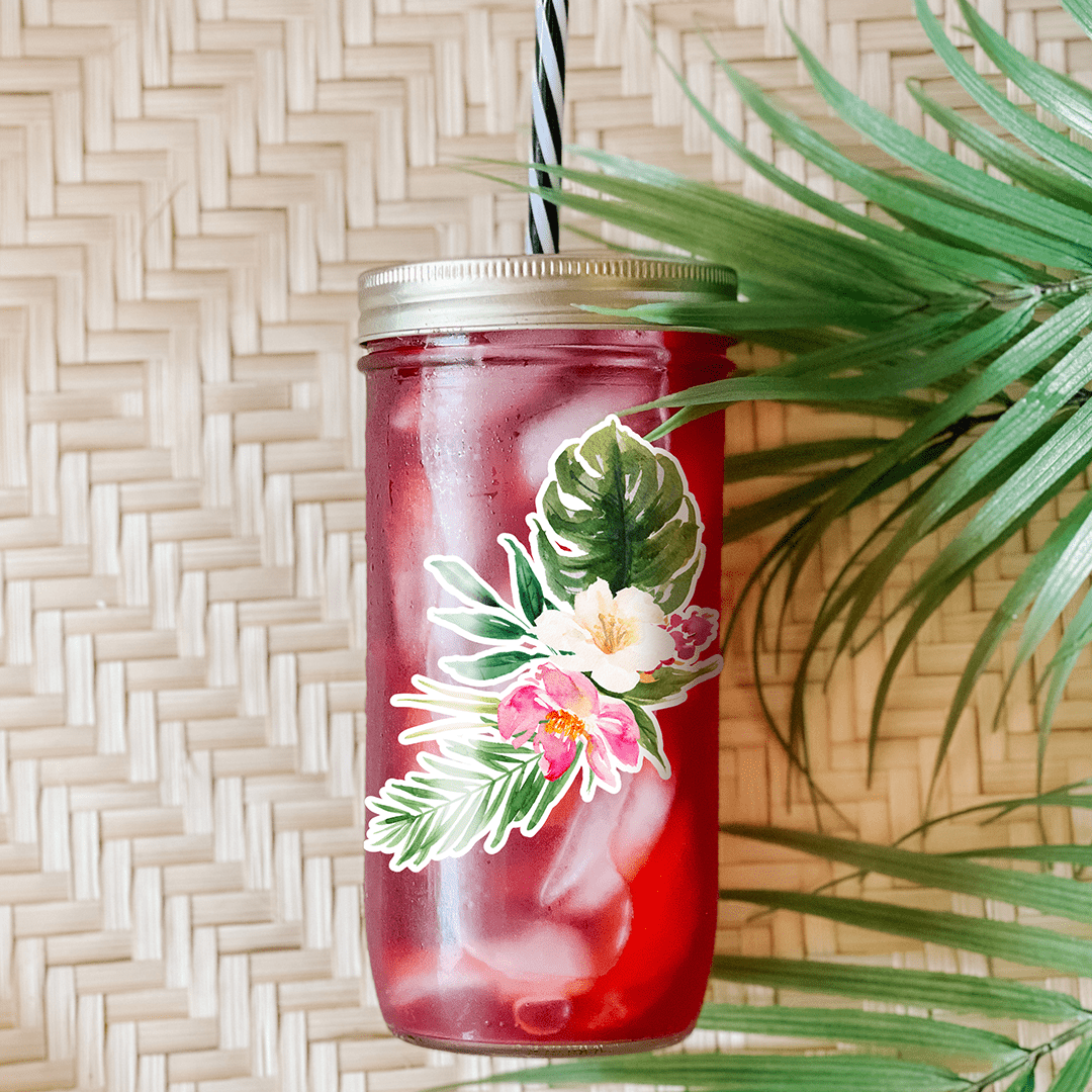 Tumbler with a red drink and a graphic sticker that has white and pink hibiscus flowers. Photographed as a flat lay in a winnowing tray and palm leaves.