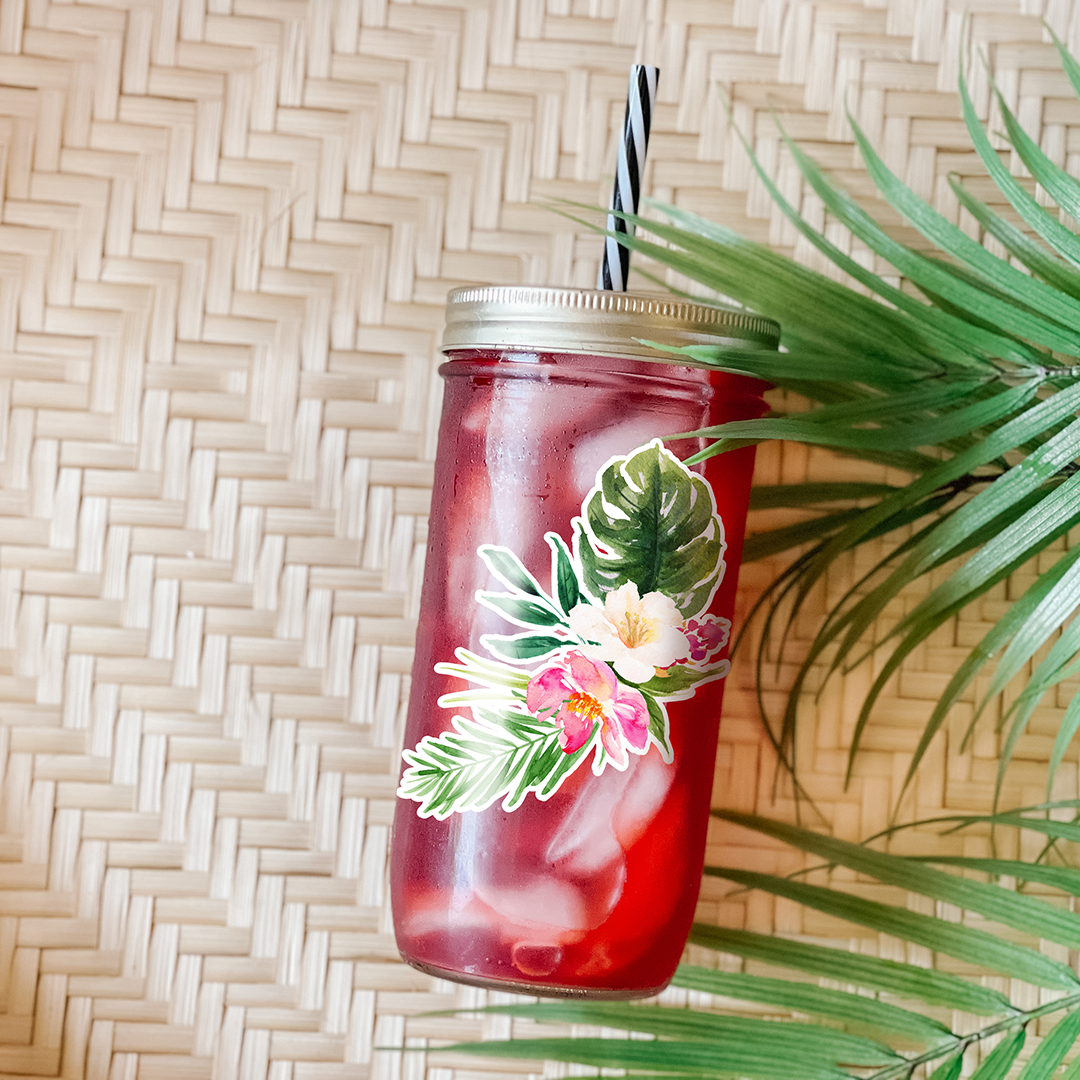 Tumbler with a red drink and a graphic sticker that has white and pink hibiscus flowers. Photographed as a flat lay in a winnowing tray and palm leaves.