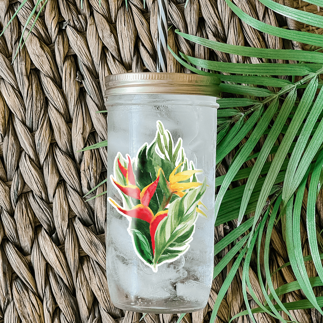 Tumbler with water and a sticker that has a graphic print of heliconia tropical flowers against palm leaves. Photographed as a flat lay in a weave mat with palm leaves.