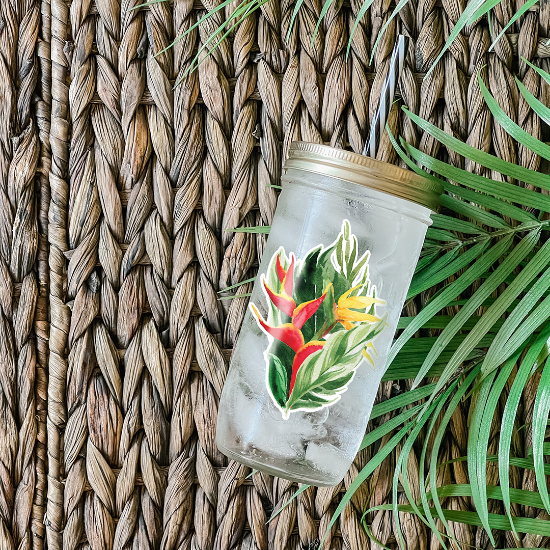 Tumbler with water and a sticker that has a graphic print of heliconia tropical flowers against palm leaves. Photographed as a flat lay in a weave mat with palm leaves.