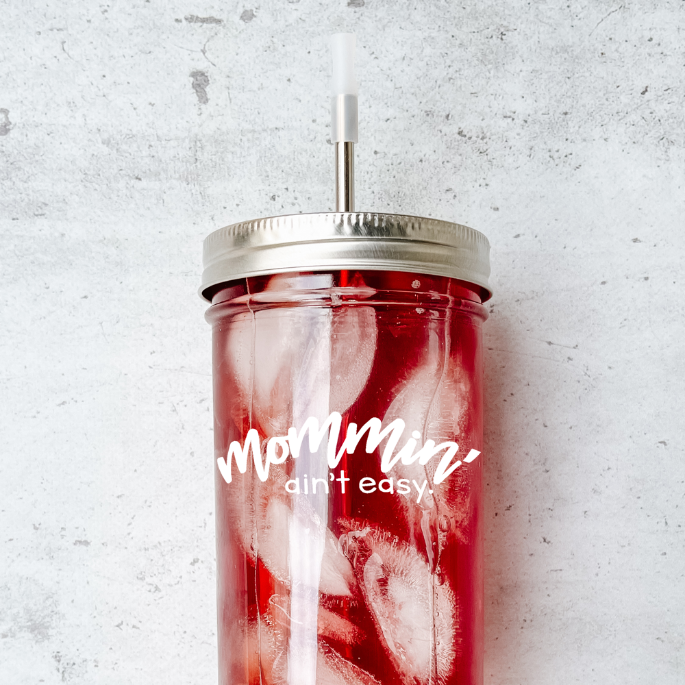 Mason jar drinking glass with a pink iced drink it it, with the phrase "mommin' ain't easy" in white on it.  It has a silver metal straw and a clear silicone straw tip. Against a marble background