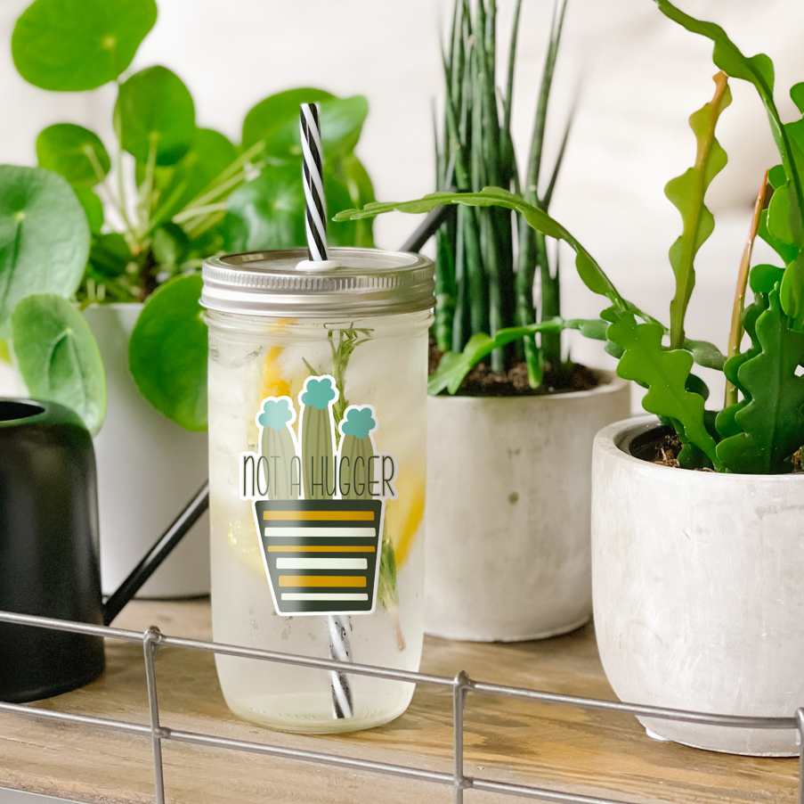 Tumbler with water inside and a sticker of cactuses on a striped pot printed on it. There is also a print that reads "Not a Hugger." Tumbler is photographed standing on a wooden table with some potted plants surrounding it.