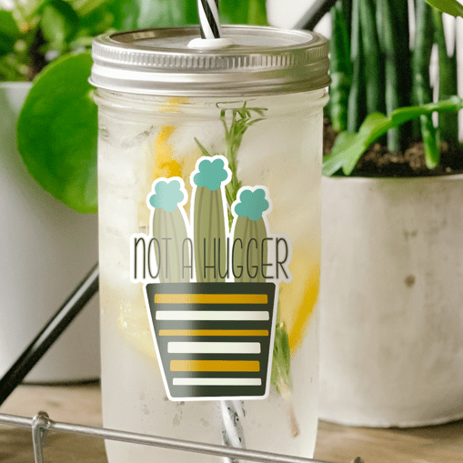 Tumbler with water inside and a sticker of cactuses on a striped pot printed on it. There is also a print that reads "Not a Hugger." Tumbler is photographed standing on a wooden table with some potted plants surrounding it.