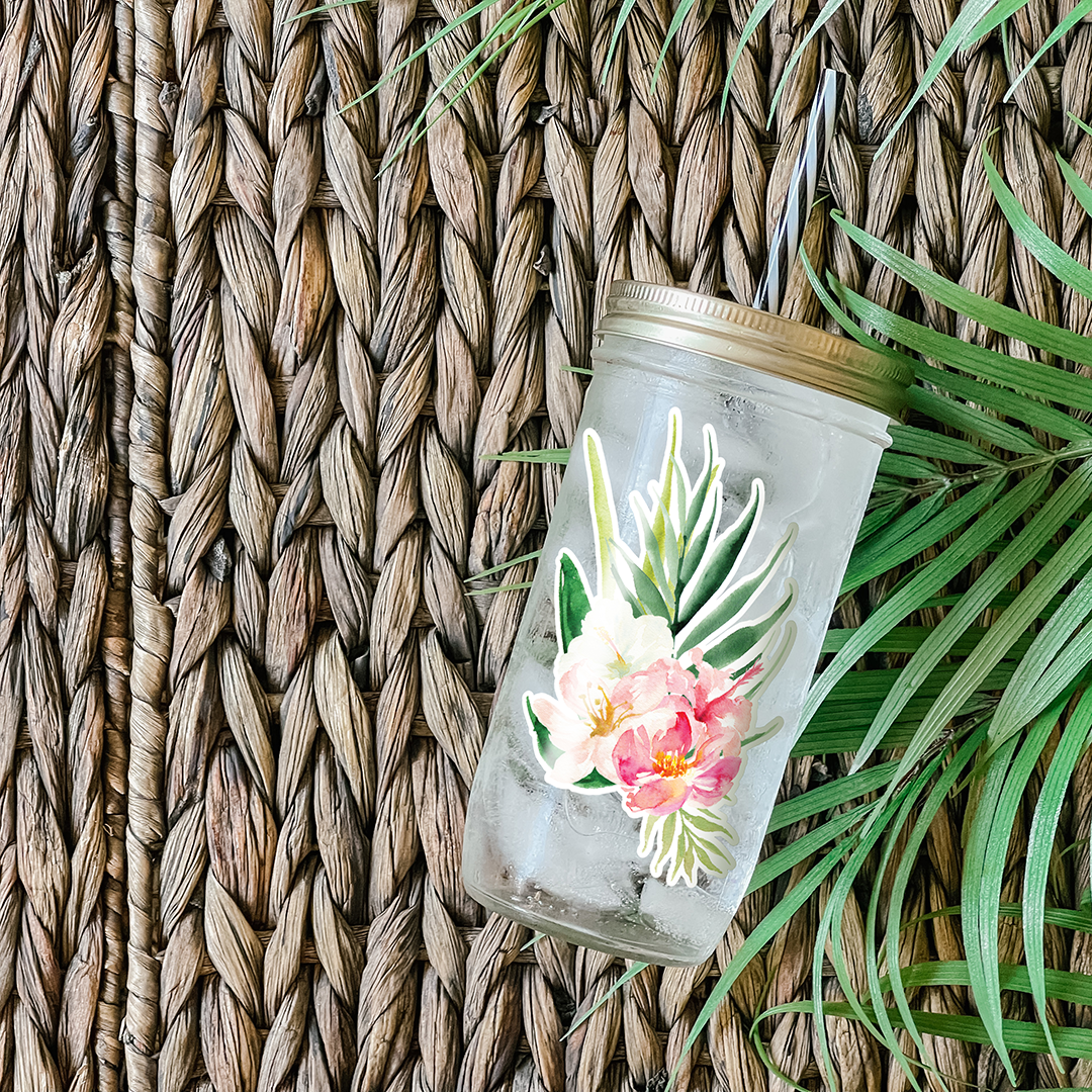 Tumbler with water and a graphic sticker that has white and pink hibiscus flowers. Photographed as a flat lay in a weave mat and palm leaves.