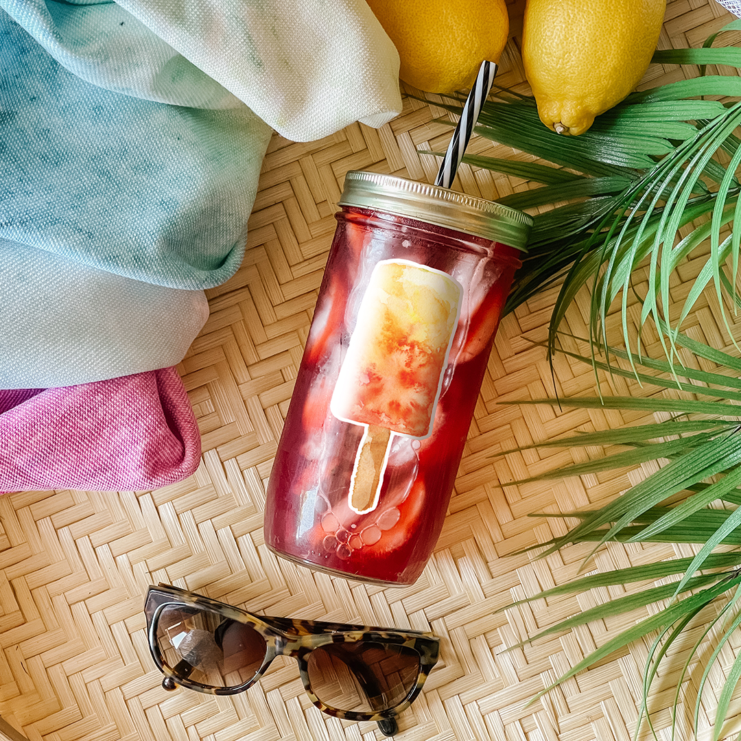 Tumbler with red drink and a single papaya-mango popsicle graphic sticker. Photographed as a flat lay in a winnowing tray with lemons, a scarf, sunglasses, and palm leaves.
