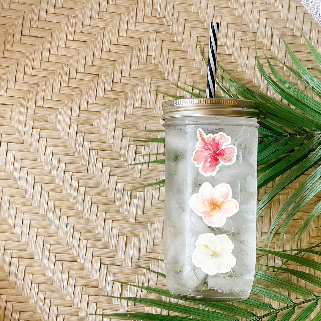 Tumbler with water and a sticker that has a pink, peach, and white hibiscus flowers. Photographed as a flat lay in a winnowing tray with palm leaves.