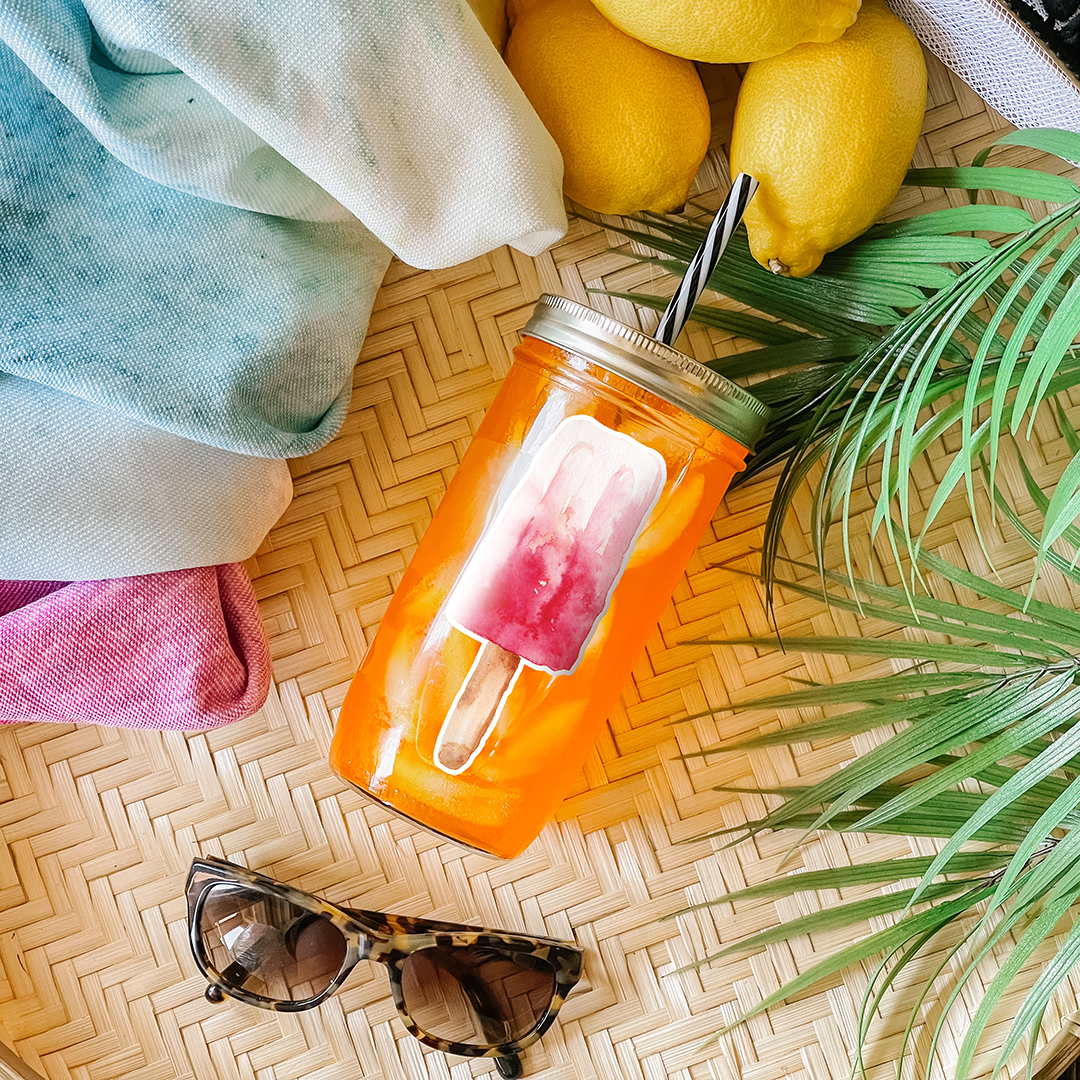 Tumbler with orange drink and a single popsicle in shades of pink graphic sticker. Photographed as a flat lay in a winnowing tray with lemons, a scarf, sunglasses, and palm leaves.