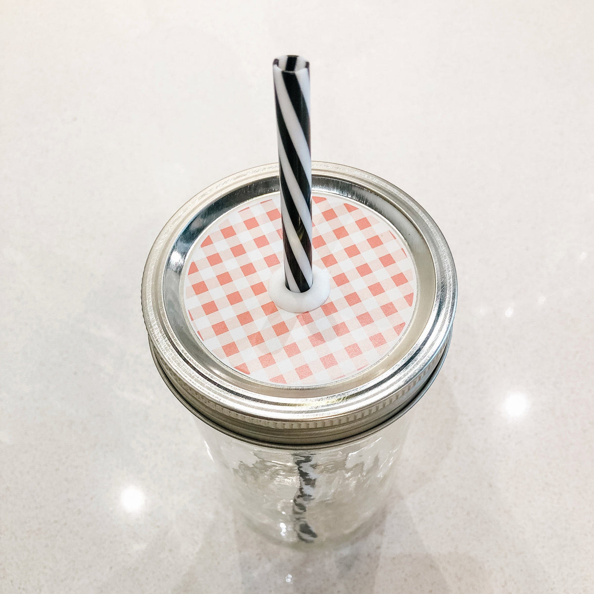 A silver lid on a regular mouth mason jar with white and peach checkered straw lid on top. It has a black and white striped straw and displayed on a white counter.