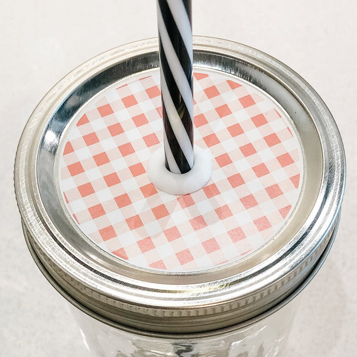 A closer look of a white and peach checkered straw lid on a silver mason jar cover. It has a black and white striped straw and displayed on a white counter.