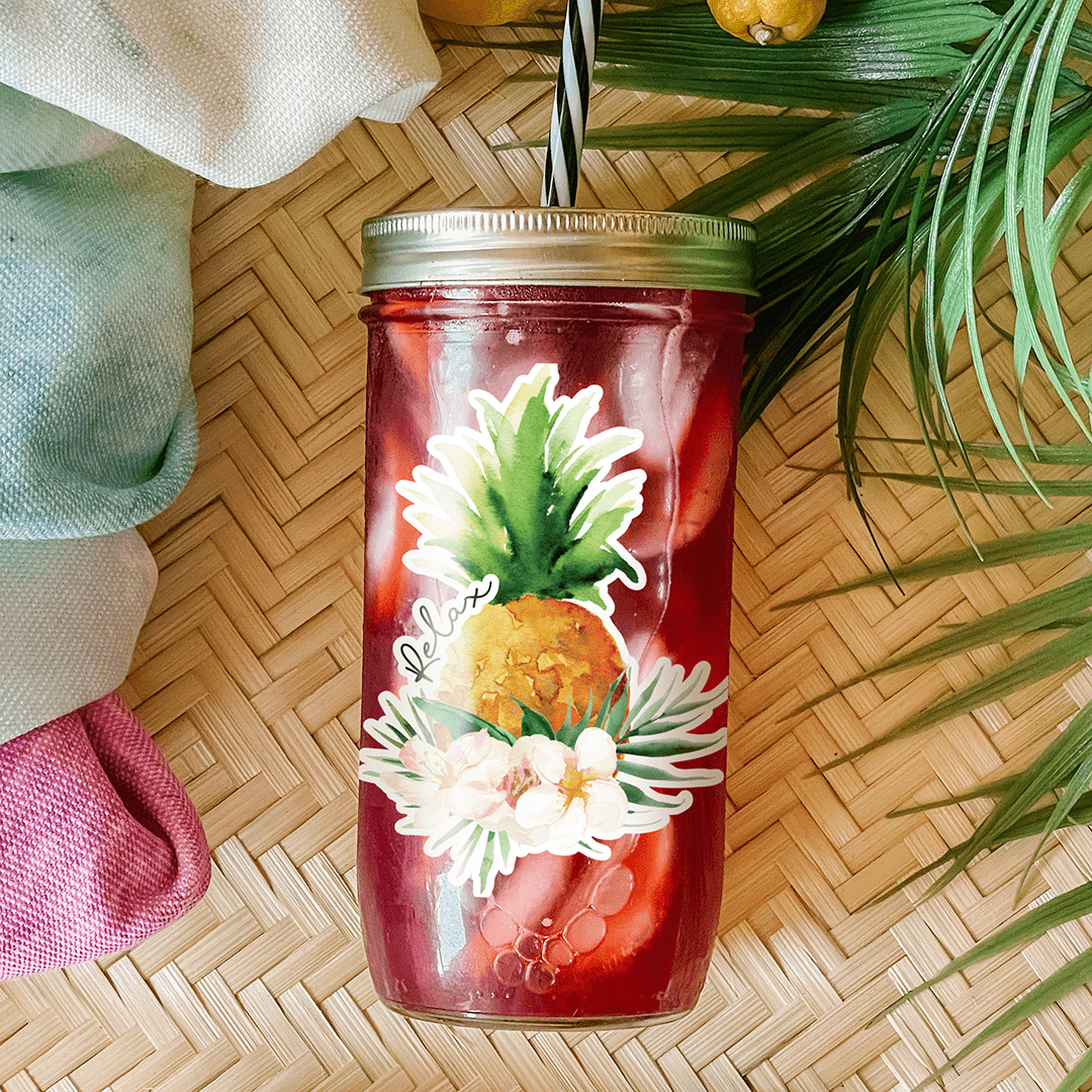 Tumbler with a red drink and a sticker that reads "Relax" in small print with a pineapple and some tropical graphic details (hibiscus flowers and palm leaves). Photographed as a flat lay in a winnowing tray with lemons, a scarf, sunglasses, and palm leaves.