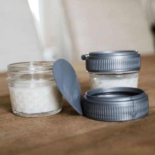 Two glass reusable mason jars filled with milk sit on a wooden counter. On one jar is a plastic mason jar ring and another ring sits on the counter