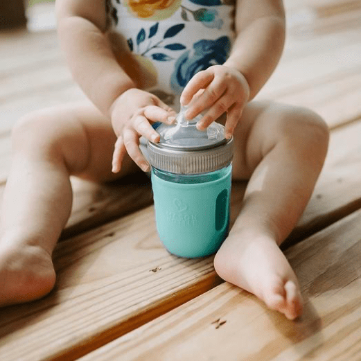 A small child sits behind a glass reusable mason jar bottle with an aqua silicone sleeve, a plastic mason jar ring and a rubber bottle top
