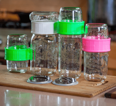 Four different sizes of to go jars with spill proof lids in different colours of green, white and pink. They sit on a cutting board on a kitchen counter