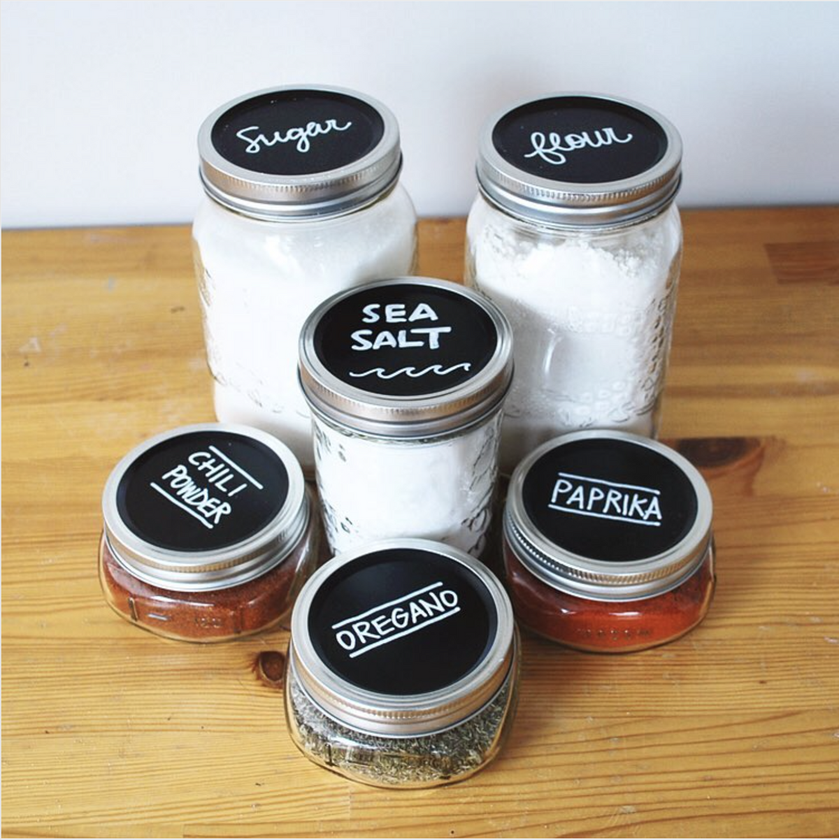 6 various wide mouth mason jars with MasonTops Chalkboard lids on top.  Various pantry ingredients are in the jars and the lid label reflects the ingredients.