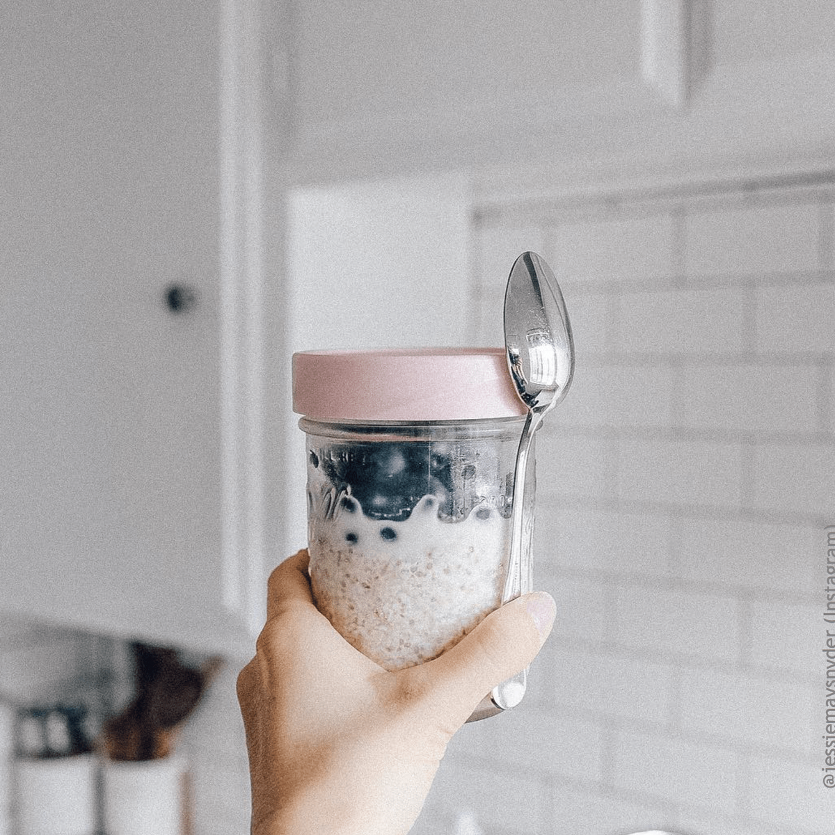 a hand holds a glass reusable mason jar with a pink lid and a spoon on a white kitchen background, the jar is filled with fruit oats and milk