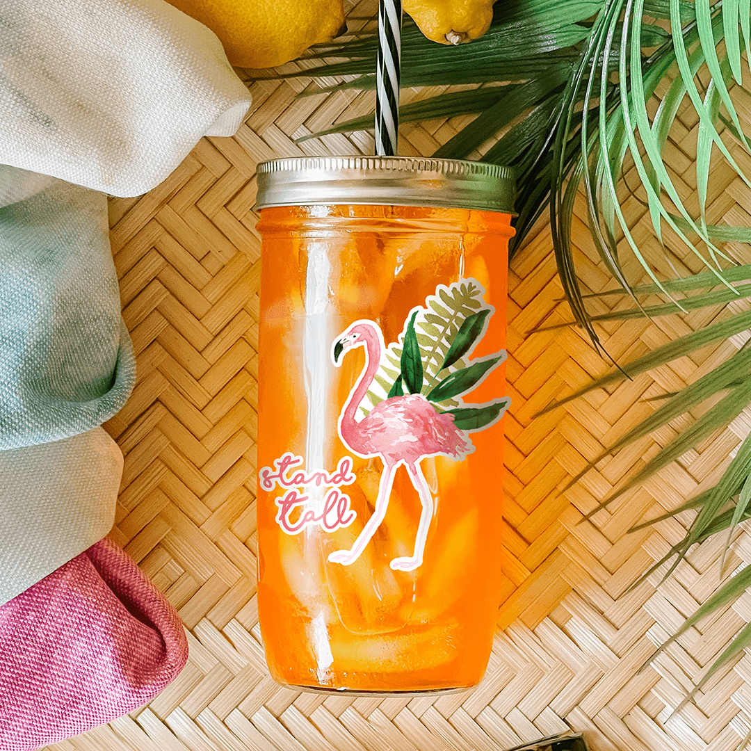 Tumbler with orange drink and a sticker that reads "Stand Tall" with a flamingo and tropical palm leaves graphic. Photographed as a flat lay in a winnowing tray with lemons, a scarf, sunglasses, and palm leaves.