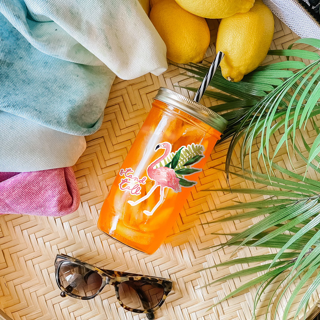 Tumbler with orange drink and a sticker that reads &quot;Stand Tall&quot; with a flamingo and tropical palm leaves graphic. Photographed as a flat lay in a winnowing tray with lemons, a scarf, sunglasses, and palm leaves.