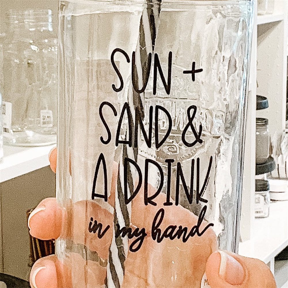 A close up of a Photo of a mason jar tumblr with a sticker text that says &quot;Sun + Sand &amp; a Drink in my hand&quot;. It has gold lid with black and white straw.A photo of a mason jar tumbler with a sticker text that says &quot;Sun + Sand &amp; A Drink in my hand&quot;