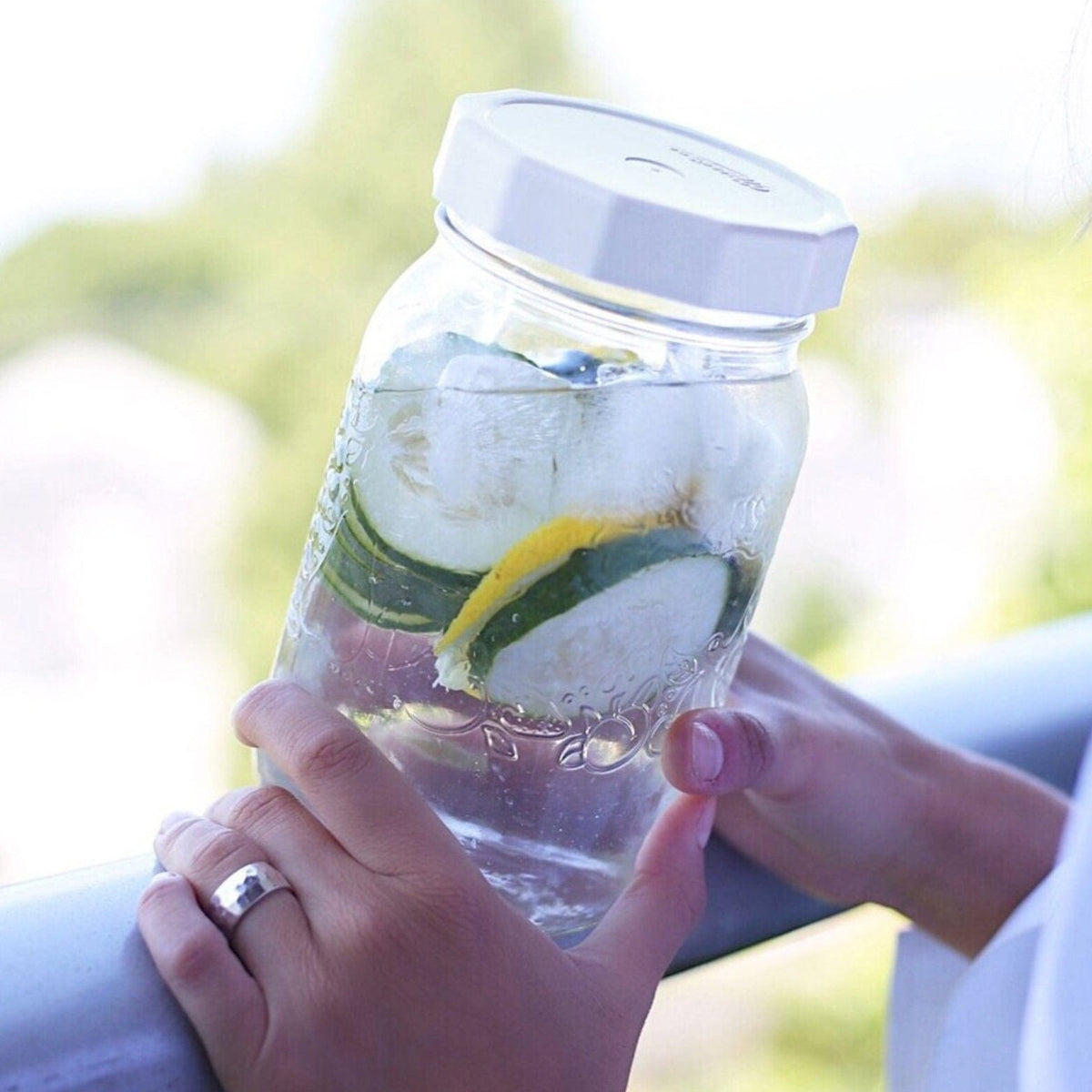 Woman is outdoors holding a quart size mason jar with lemon cucumber water in it with a white MasonTops tough top on the jar.