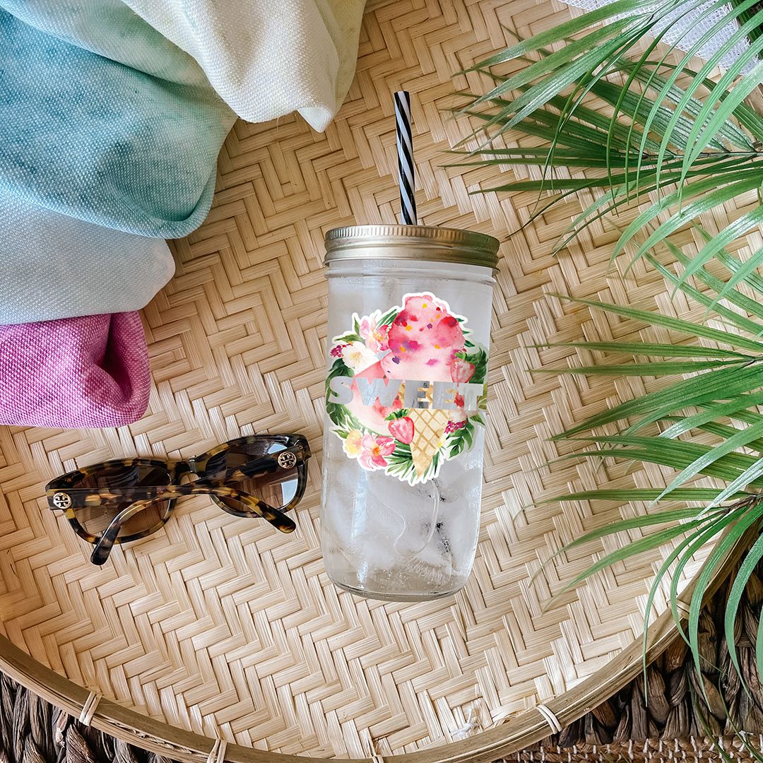Tumbler with water and a graphic sticker that reads "Sweet" in silver print surrounded by tropical details graphics. Photographed as a flat lay in a winnowing tray with a scarf, sunglasses, and palm leaves.