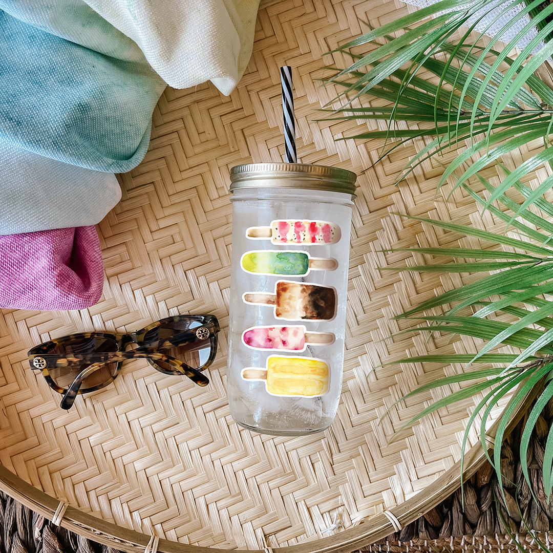 Tumbler with water and multicolored popsicle graphic stickers. Photographed as a flat lay in a winnowing tray with a scarf, sunglasses, and palm leaves.