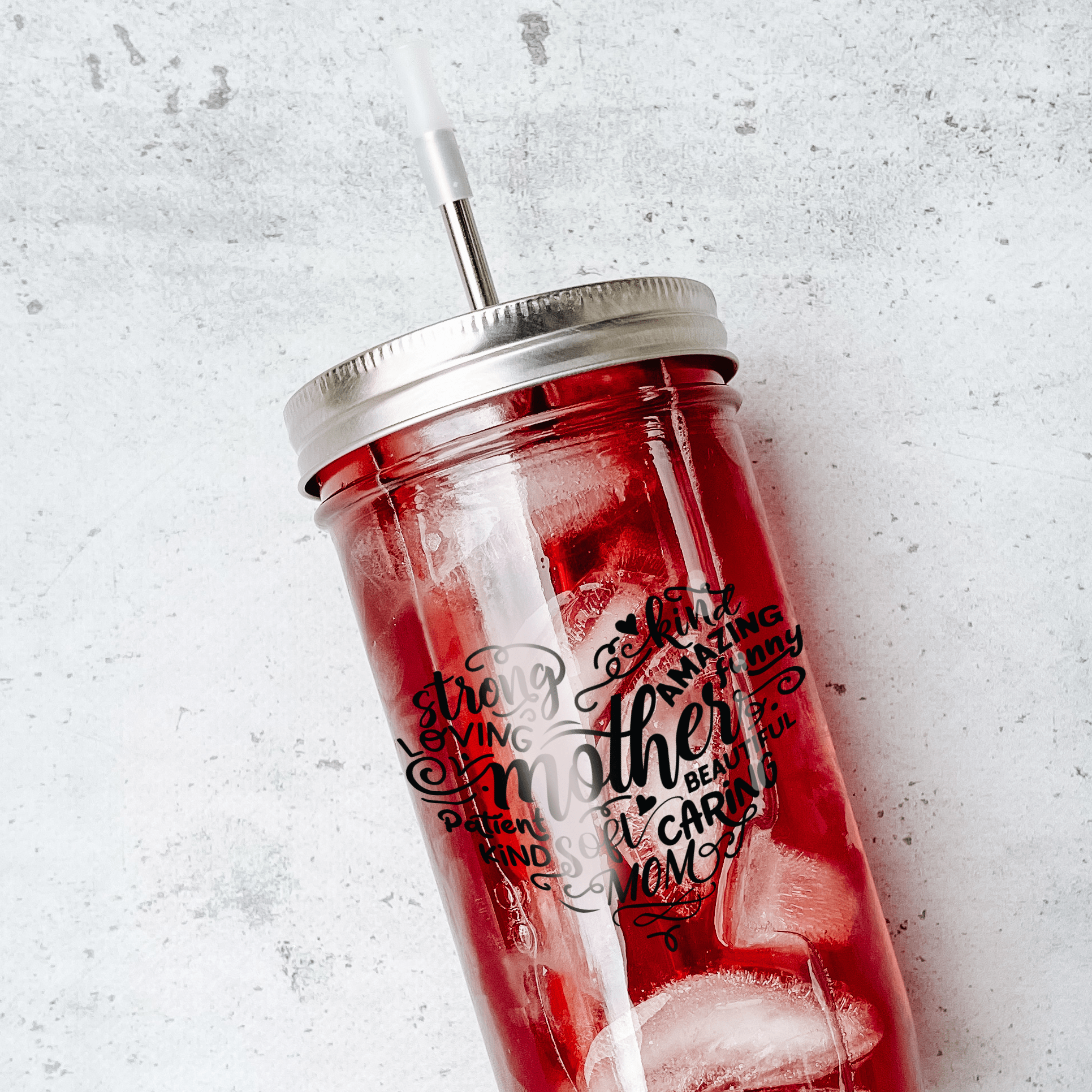 Mason jar tumbler with red drink and a silver straw and lid with print that reads "mother" in black surrounded by descriptive words for mother in black too. Against marble background.