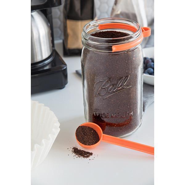 32 oz reusable glass mason jar sits on a kitchen counter containing coffee grounds. To the side sits an orange coffee scoop and on the jar is an orange coffee clip