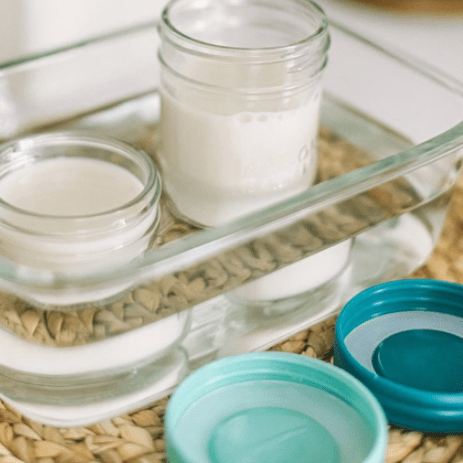 8oz and 4oz mason jar bottles with milk stored. Both are dipped in warm water in a rectangular glass container. The container is placed on top of a native tray with Ocean and Aqua colored lids beside it. Photographed against a white countertop.