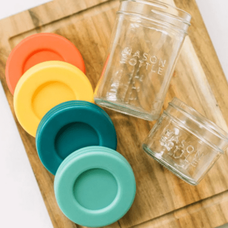 Empty 8oz and 4oz mason jar bottles and plastic lids in Paprika, Mango, Ocean, and Aqua. Photographed as a flat lay in a chopping board on a white countertop.