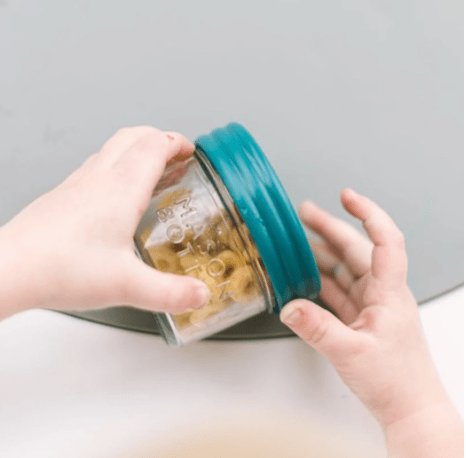 A child holding a 4oz mason jar bottle with Ocean colored lid and snacks inside. Photographed against a white countertop with a gray placemat.