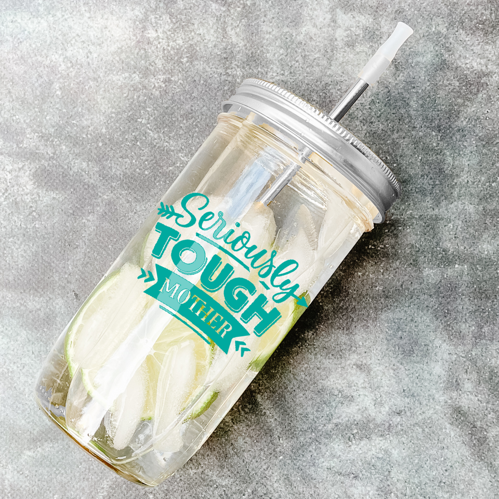 Mason jar tumbler with citrus in water drink and a silver straw and lid with print that reads "Seriously Tough Mother" in black. Against marble background